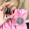 A hand holds a black key with a tag and a small silver chain, next to a clear plastic CD keychain.
