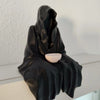 A black resin statue of a draped ghost figure holding a small, light-colored tea cup.