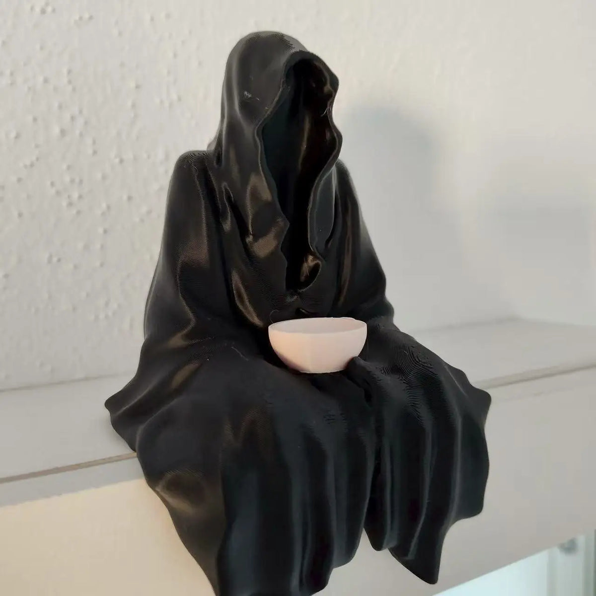 A black resin statue of a draped ghost figure holding a small, light-colored tea cup.