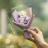 A hand holds a handmade crochet flower bouquet featuring purple and white flowers in a pink paper wrapping.