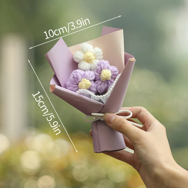 A hand holds a handmade crochet flower bouquet featuring purple and white flowers in a pink paper wrapping.