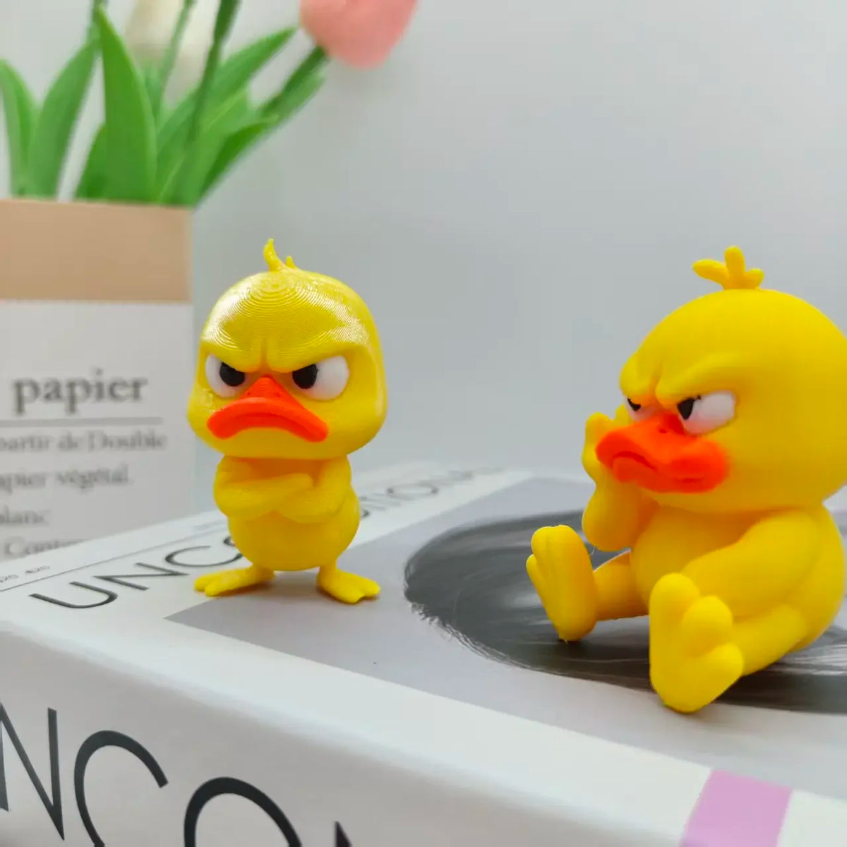 A yellow 3D printed angry duck figurine with orange beak sitting on a white box.