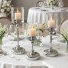 A silver metal pillar candle holder with a white candle is displayed on a white tablecloth for wedding or home decor.