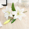 Realistic silk lily flower stem with three white blooms and green leaves on a wooden surface.