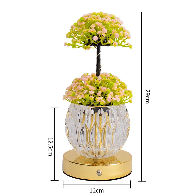 A decorative plant arrangement featuring pink and green flowers on a black stem, displayed in a clear glass pot on a gold bas