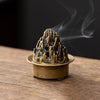 A vintage brass incense burner holder with a textured top is emitting smoke on a wooden surface.