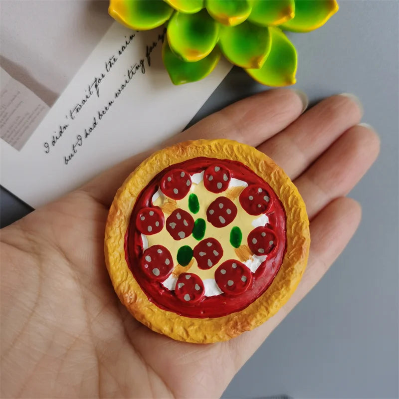 A hand holds a round pizza-shaped food magnet with melted cheese and toppings against a light background.