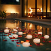 Floating candles illuminate a tranquil pool at dusk with reflections on the water.