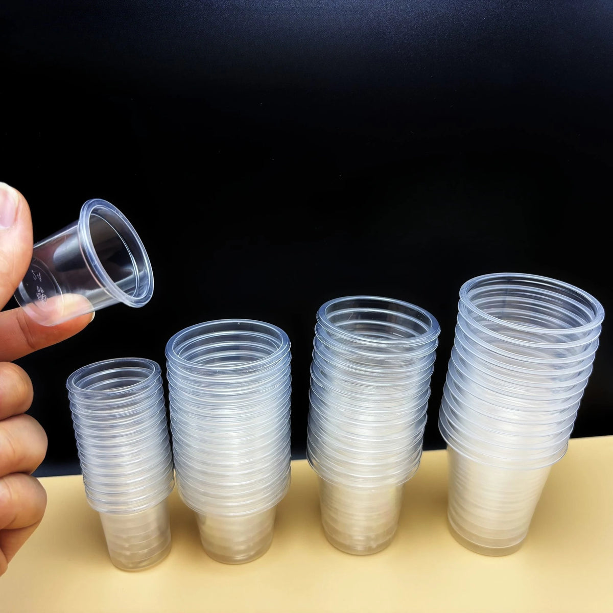 A hand holds a stack of clear disposable mini tasting cups on a light surface against a black background.