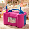 A pink and blue handheld electric balloon pump sits on a light rug near a bed with pastel decorations.