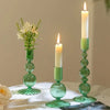 A set of three groovy wavy glass taper candle holders with green candles and flowers on a white surface.