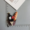 A set of playful food-shaped fridge magnets including a bottle, a strawberry, and a croissant, arranged next to a ruler.