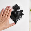 A hand holds a black resin heart decoration shaped like flames and leaves on a white background.