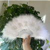 A white feather hand fan decorated with rhinestone letters spelling BRIDE, held by a person against a green leafy background.