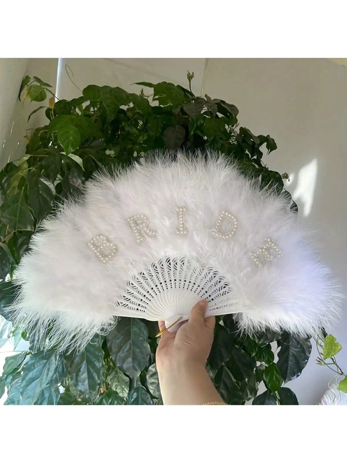 A white feather hand fan decorated with rhinestone letters spelling BRIDE, held by a person against a green leafy background.