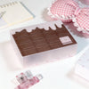 Una caja rectangular de pegatinas o cartas de chocolate marrón junto a tela decorativa rosa y un pequeño plástico
