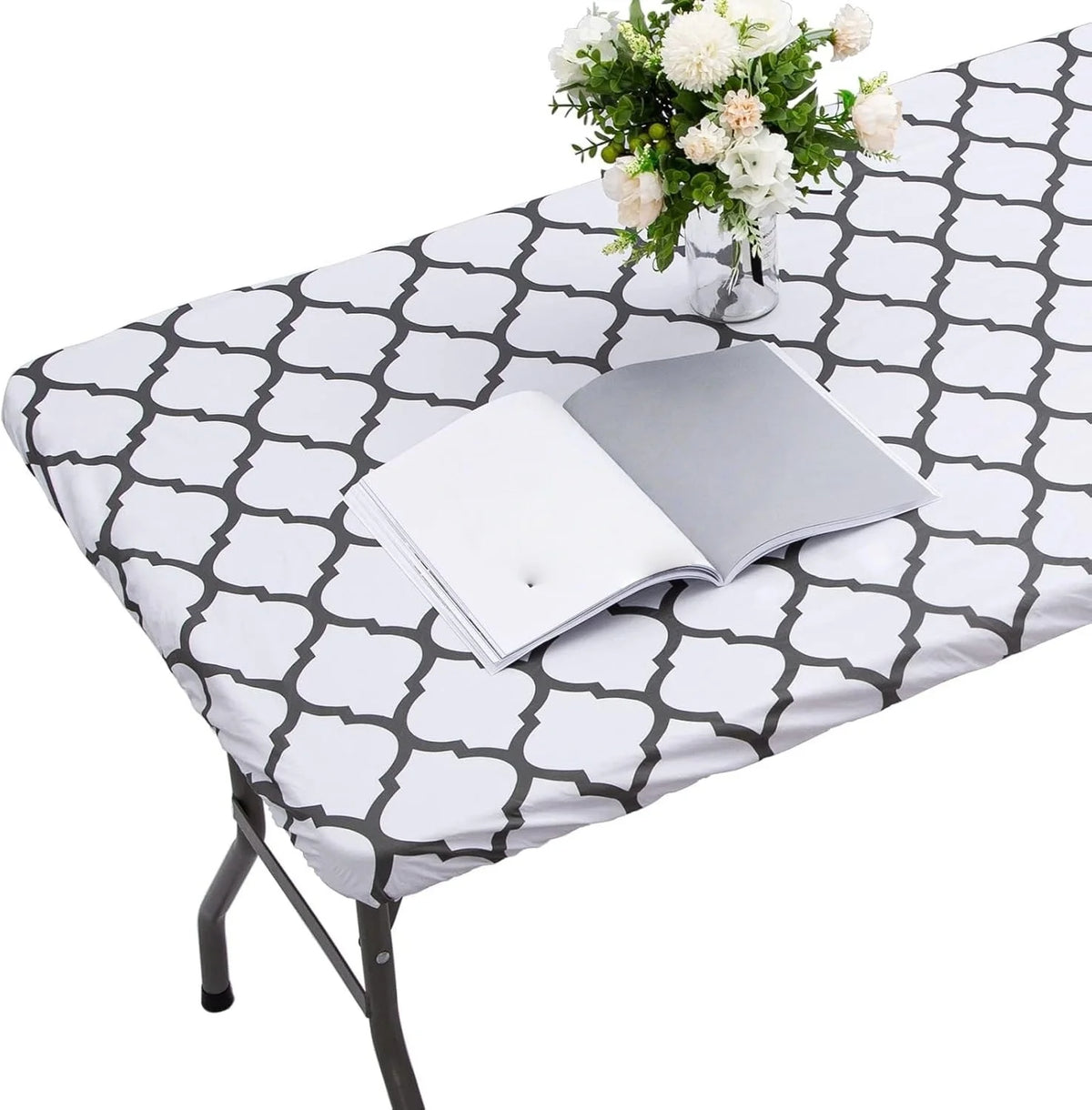 A white and gray patterned elastic fitted rectangle tablecloth is displayed on a table, with a stack of white papers and a va