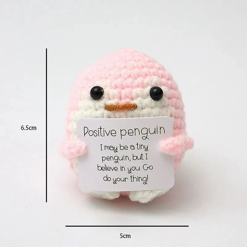 A handmade crochet pink penguin doll with an inspirational card, shown next to a scale.