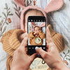 A baby is held in a knitted hat and is looking at a smartphone displaying a personalized newborn hospital keepsake sign.