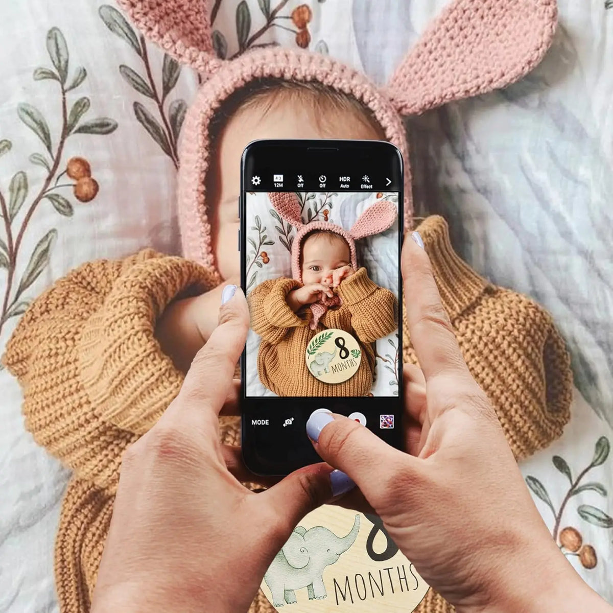 A baby is held in a knitted hat and is looking at a smartphone displaying a personalized newborn hospital keepsake sign.
