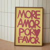 A framed canvas print with pink text that reads MORE AMOR POR FAVOR on a light yellow background.