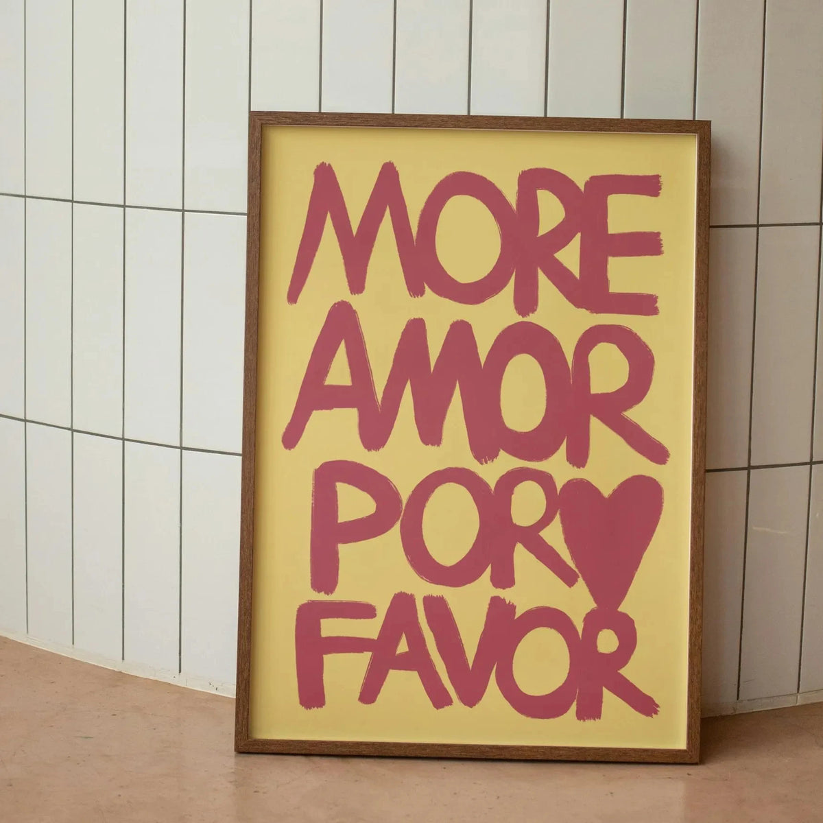 A framed canvas print with pink text that reads MORE AMOR POR FAVOR on a light yellow background.