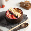 A dark coconut shell serving bowl filled with sliced bananas, strawberries, and seeds on a gray linen cloth.
