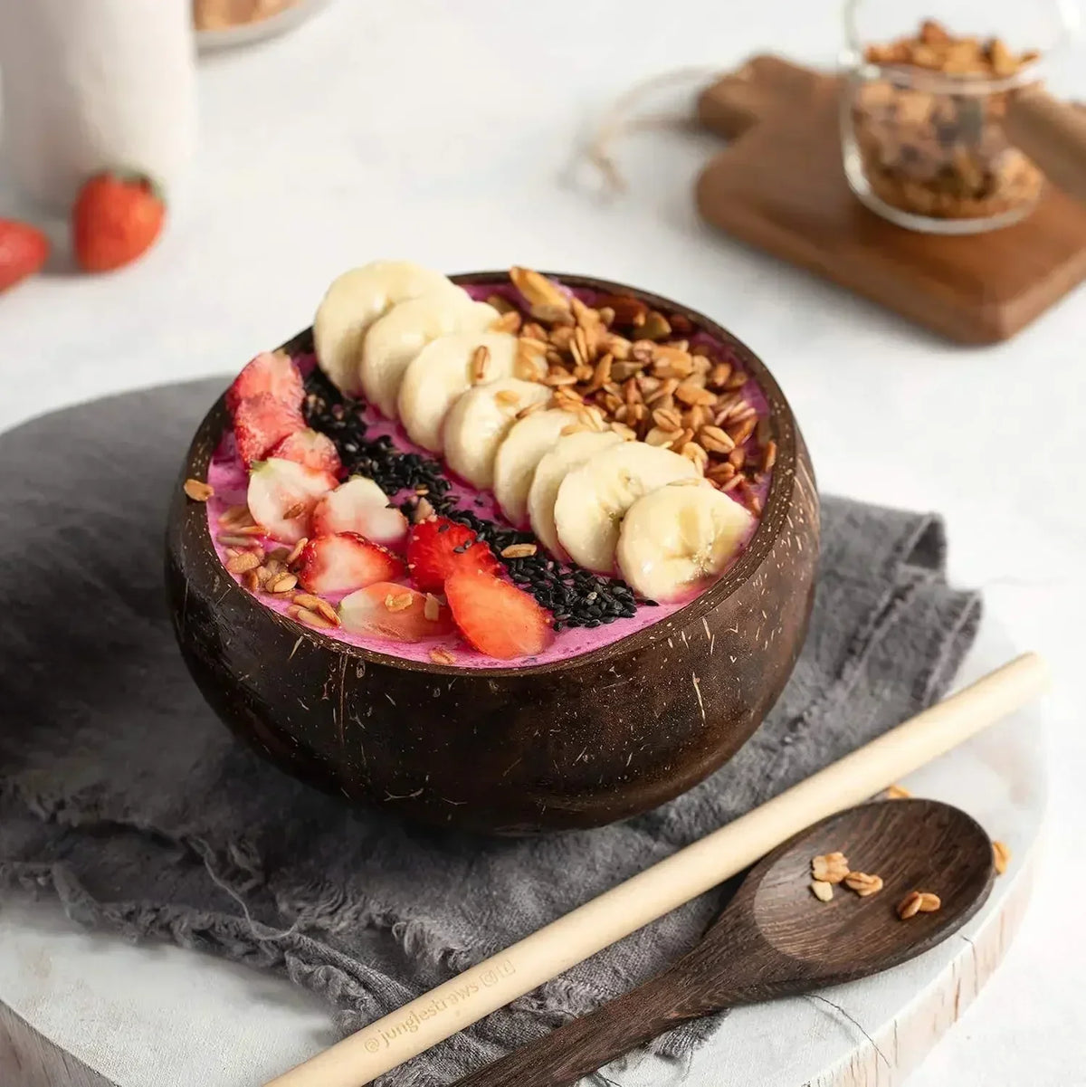 A dark coconut shell serving bowl filled with sliced bananas, strawberries, and seeds on a gray linen cloth.