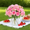 A bouquet of pink artificial silk peony flowers in a glass vase on a white cloth outdoors.