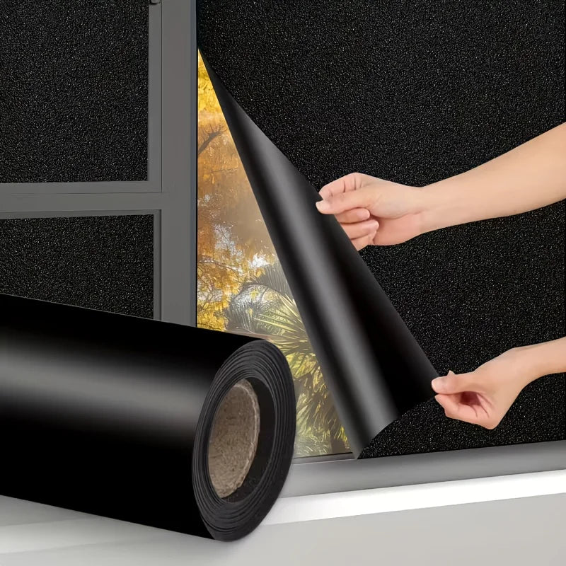 A hand is pulling a roll of black privacy window film across a window pane.