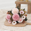 A bouquet of pink and white silk flowers including roses and hydrangeas arranged on a wooden board.