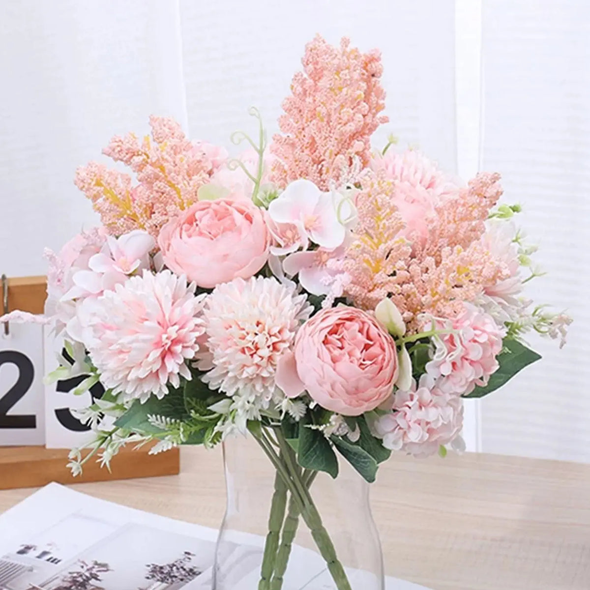 A bouquet of artificial silk peony flowers in shades of pink and peach arranged in a clear glass vase.