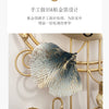 A close-up of a decorative metal ginkgo leaf wall clock with gold accents, featuring textured silver and blue tones.