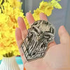 A hand holds a black and white metal skull emblem decal against a background of yellow flowers.