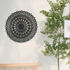 Decoración de pared de mandala de madera negra colgada en una pared blanca junto a una rama verde y frondosa.