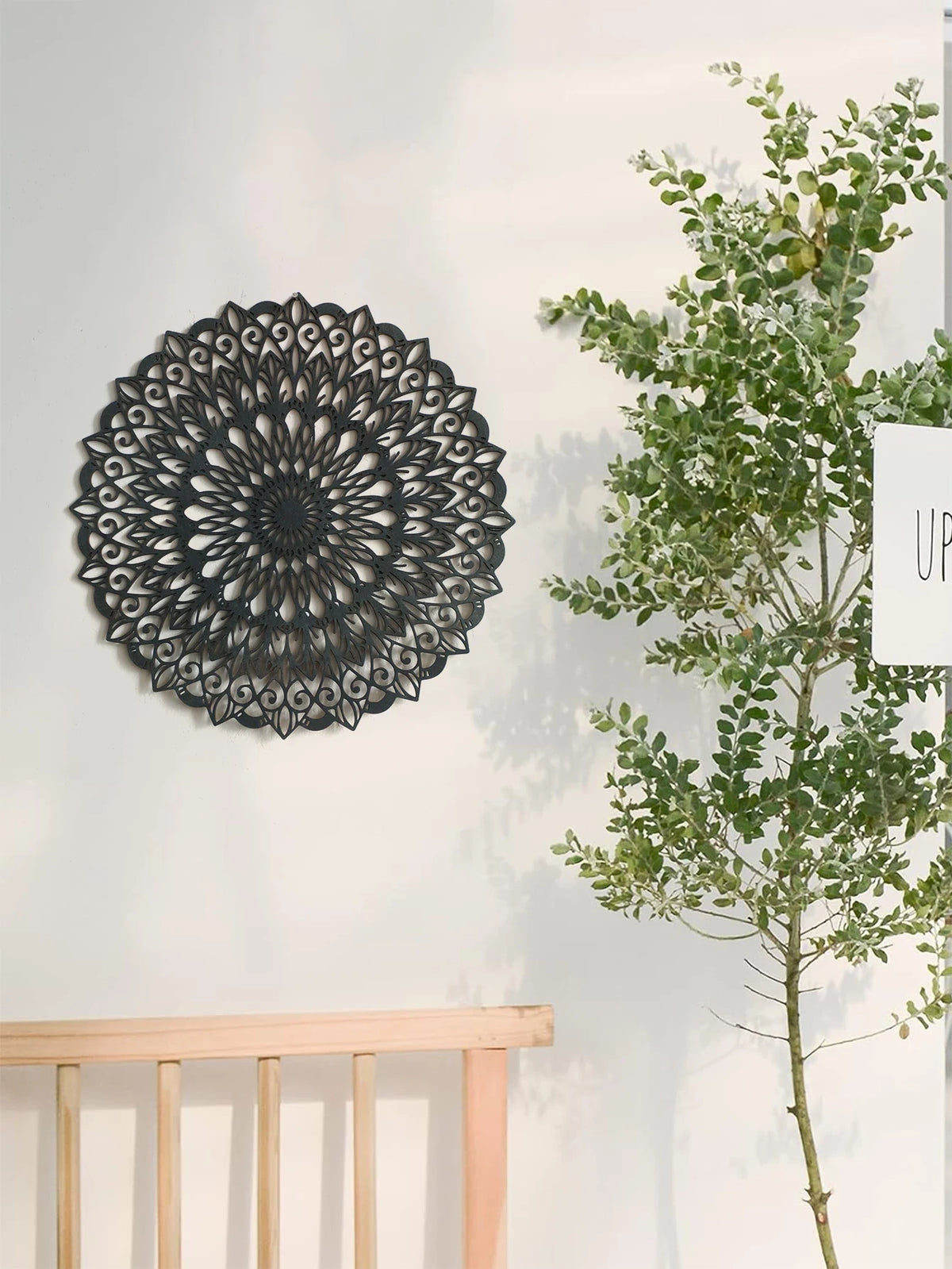 Decoración de pared de mandala de madera negra colgada en una pared blanca junto a una rama verde y frondosa.