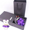 Una caja de regalo gris oscuro con un ramo de rosas moradas y una figura de oso blanco con una cinta de 'te amo'.