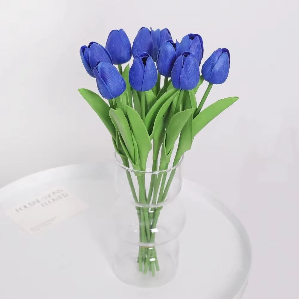 A bouquet of vibrant blue artificial tulip flowers in a clear glass vase on a white surface.