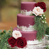 A three-tiered purple cake decorated with pink and deep red artificial roses and green foliage on a silver platter.
