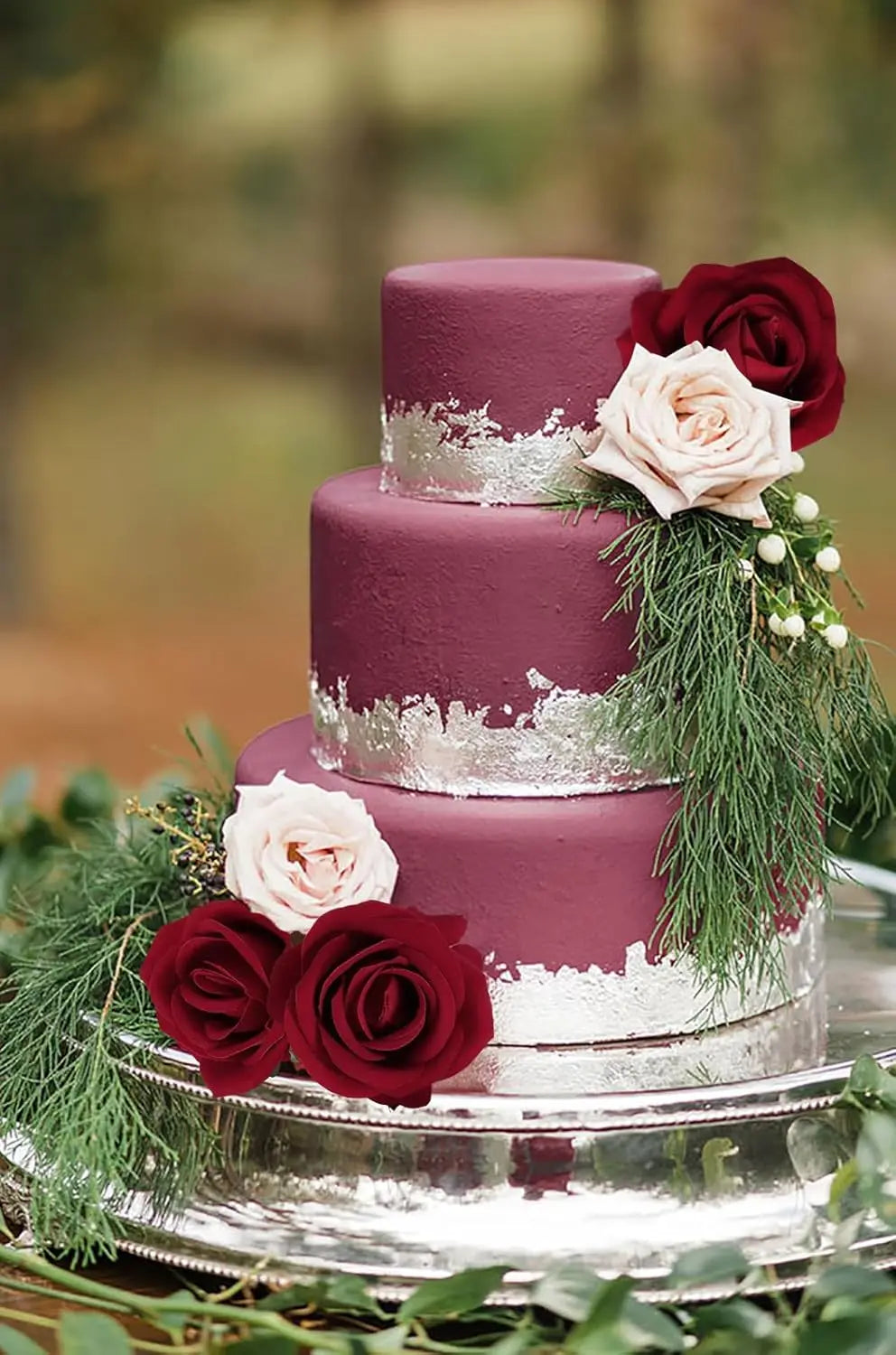 A three-tiered purple cake decorated with pink and deep red artificial roses and green foliage on a silver platter.