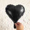 A black heart-shaped foil balloon being held by a hand on a white surface.