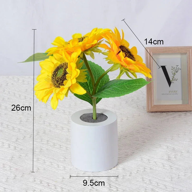 A bright yellow sunflower plant in a white pot with leaves and flowers, shown next to a framed picture.