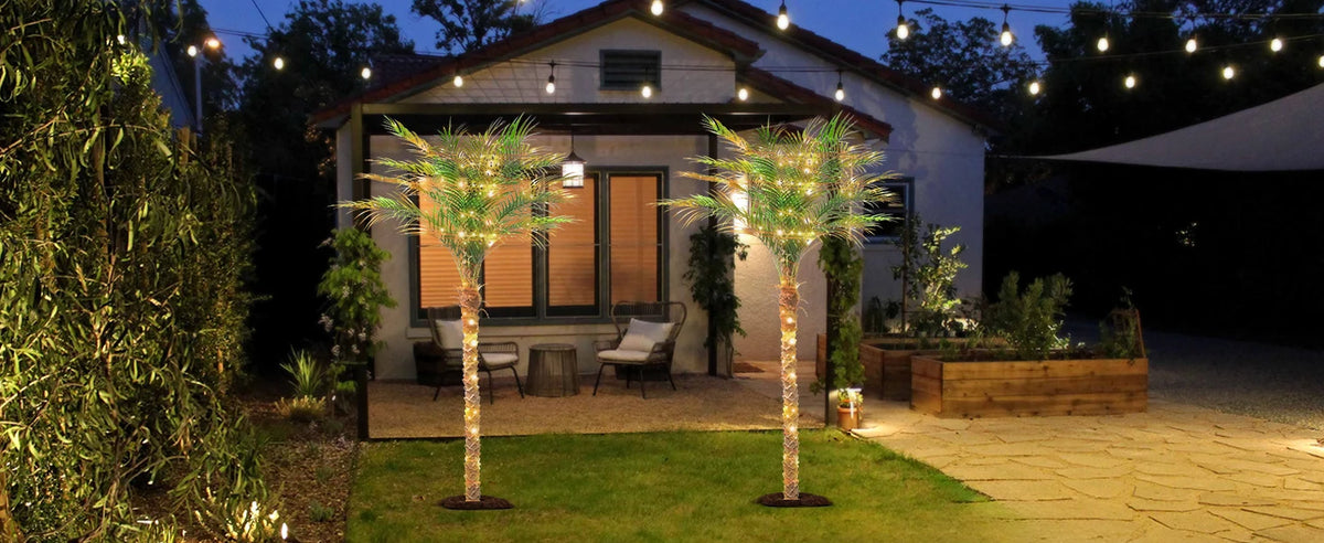 An artificial golden palm tree stands in a lawn next to a house at dusk, illuminated by string lights.