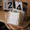 Une boîte d'organisation de bureau en acrylique transparent contient une pile de papiers et un calendrier, sur un fond en boi