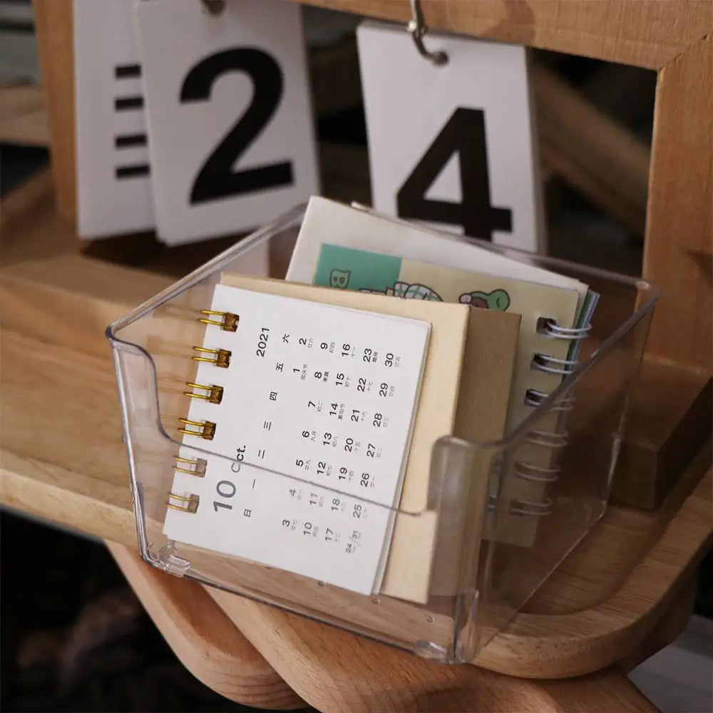 Une boîte d'organisation de bureau en acrylique transparent contient une pile de papiers et un calendrier, sur un fond en boi
