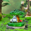 A green ceramic ornament featuring a small gray animal figure with white accents, set in a mossy outdoor scene.