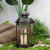 A vintage Nordic iron candle lantern with glass panes sits on mossy ground next to a large rock and greenery.