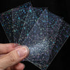 A hand holds several holographic card sleeves with a dark purple and teal star pattern.