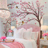 A bedroom decorated with pink cherry blossom tree wall stickers, featuring a pink upholstered bed and white linens.