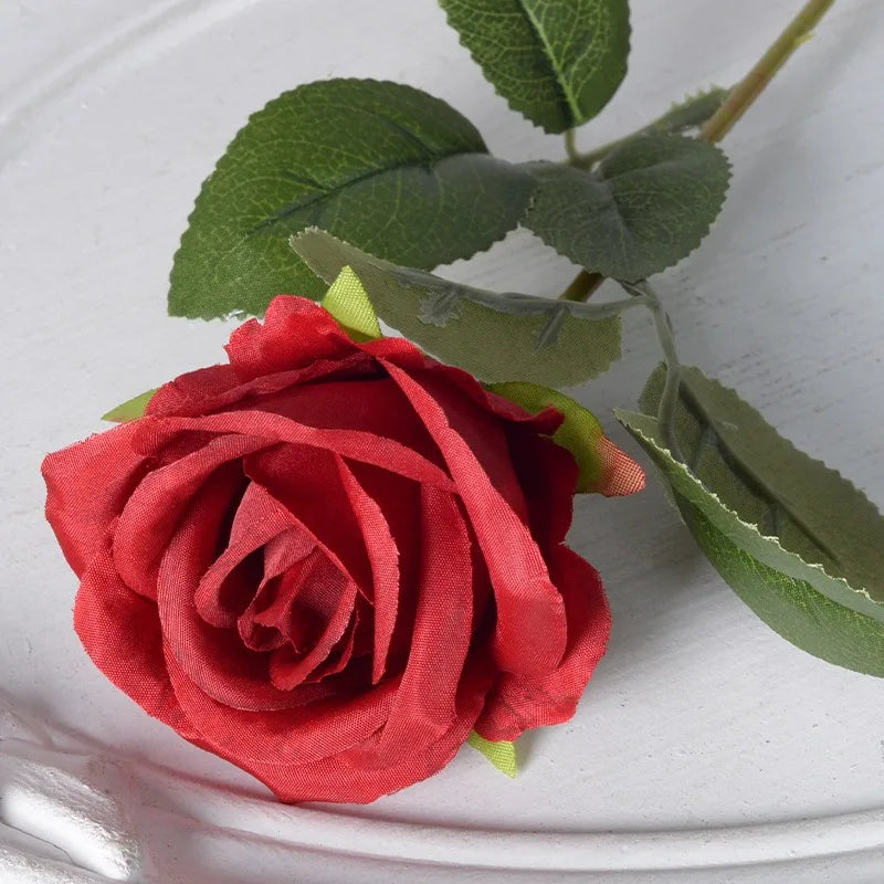 A vibrant red velvet artificial rose with green leaves on a white background.